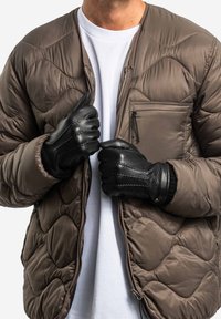 Braune gesteppte Pufferjacke mit einer Reißverschlusstasche, getragen über einem weißen T-Shirt, und schwarzen Lederhandschuhen mit strukturierten Akzenten.