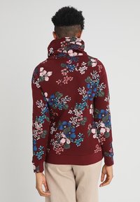 Pullover floreale marrone con collo alto, caratterizzato da un motivo di fiori blu, rosa e verdi su un tessuto morbido e testurizzato. Polsi e orlo a coste.
