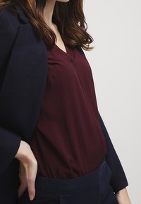 Burgundy blouse with a v-neck, paired with a navy blazer. The blouse has a smooth texture and subtle gathering at the waist.