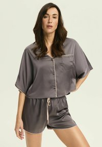Kobieta z długimi brązowymi włosami, ubrana w szary satynowy top z krótkimi rękawami z guzikami oraz dopasowane spodenki z wiązanym pasem, stojąca na tle jednolitym.