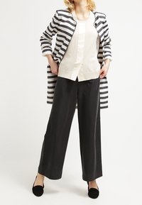 Randig svart och vit lång jacka, beige topp, svarta vida byxor, svarta platta skor. Texturerad tyg, knappstängningar, avslappnad passform.