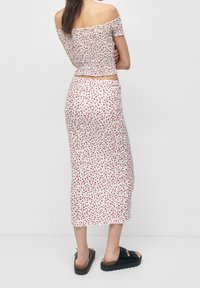 White two-piece outfit featuring an off-shoulder smocked top and a matching midi skirt, both with a red floral pattern. Worn with black sandals.