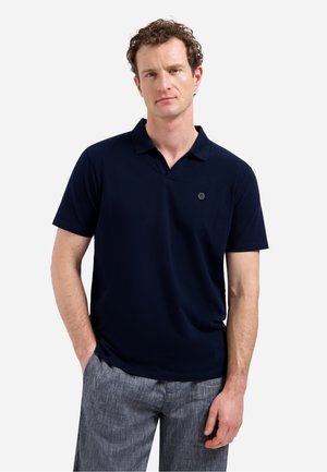 Man met krullend haar, gekleed in een marineblauw poloshirt met korte mouwen en grijze broek, met handen in de zakken, neutrale gezichtsuitdrukking, witte achtergrond.