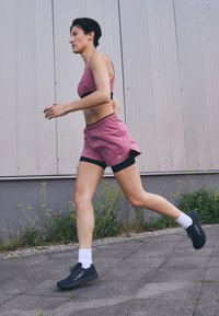 Coureuse en soutif de sport rose et shorts assortis avec des accents noirs. Chaussures de course noires, chaussettes blanches et une silhouette athlétique élancée.