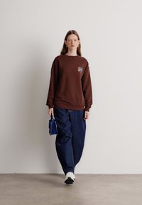 Sudadera marrón con un logotipo en la parte delantera, combinada con unos vaqueros oscuros de corte holgado. El modelo sostiene un pequeño bolso azul y lleva zapatillas blancas.