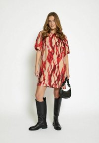 Abito rosso e beige con un motivo, maniche corte, design a A-line. Indossato con stivali neri al ginocchio e una piccola borsa nera. Tessuto morbido.