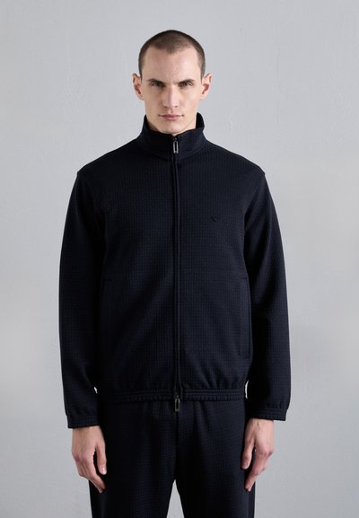 Giacca nera con zip e colletto alto, con un motivo testurizzato, tasche anteriori e polsini elasticizzati, abbinata a pantaloni coordinati.