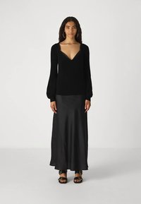 Γυναίκα που φοράει ένα μαύρο βαθύ V-ντεκολτέ πουλόβερ, μαύρη σατέν μάξι φούστα, χρυσά δαχτυλίδια και μαύρες σάντες με χρυσές αγκράφες, σε λευκό φόντο.