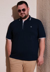 Námořnická polo košile s bílými akcenty a malým emblémem, v kombinaci s khaki kalhotami. Krátké rukávy a klasický design límečku.