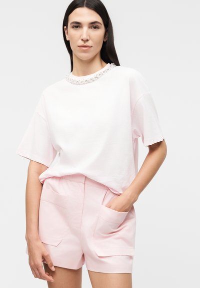 Νεαρή γυναίκα με μακριά σκούρα μαλλιά φοράει λευκό κοντομάνικο τοπ με διακοσμητικά μαργαριτάρια στο ντεκολτέ και ανοιχτό ροζ σορτς με τσέπες.