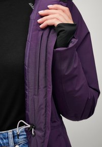 Fioletowa kurtka zewnętrzna o teksturowanym wykończeniu, z kieszeniami na zamek i sznurkiem. Nosi się ją na czarnym długim rękawie.