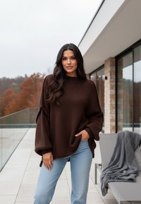Brązowy sweter dzianinowy z wysokim okrągłym dekoltem, oversize'owymi rękawami i prążkowanymi mankietami, w połączeniu z jasnoniebieskimi dżinsami na patio.