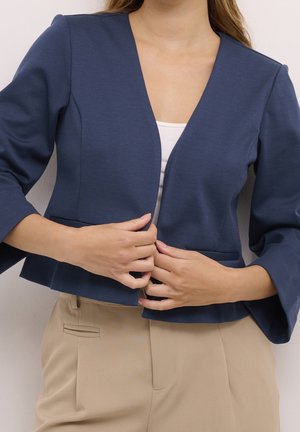 Woman wearing a navy blue blazer with three-quarter sleeves and beige pants, fastening the front of the blazer with both hands.