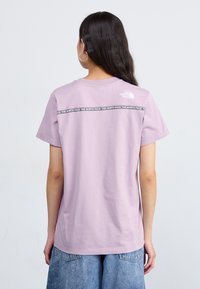 Camiseta de manga corta en color lavanda hecha de algodón. Presenta un logo blanco y texto negro repetido en la parte posterior. Corte casual con escote redondo.
