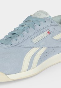 Φωτεινό μπλε σουέτ αθλητικό παπούτσι Reebok με μπλε κορδόνια, λευκές ρίγες στο πλάι και μια μικρή σημαία του Ηνωμένου Βασιλείου κοντά στο λογότυπο της Reebok.