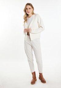 Crème gebreide cardigan met V-hals en geribde zoom, gedragen over een wit topje en gecombineerd met lichtbeige broek en bruine enkellaarzen.