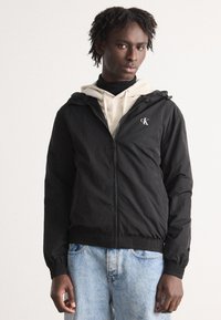 Črna zip-up jakna s kapuco, mehke teksture, z izvezenim logotipom na levem prsnem delu, v kombinaciji s svetlo bež kapuco in svetlim jeansom.