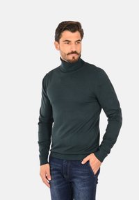 Maglione a collo alto verde scuro in una fine trama, con polsini e orlo aderenti, abbinato a jeans in denim blu.
