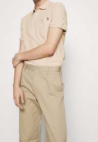 Béžové polo tričko s kapsou, zdobené malým červeným a modrým pruhem. Spárováno se světle béžovými kalhotami, obojí z hladkého materiálu.