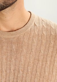 Beige stickad tröja med ribbad rund halsringning, prydd med ett texturerat kabelmönster över hela ytan, i ett mjukt och lätt tyg.