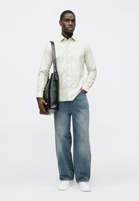 Ανοιχτό μπεζ πουκάμισο με κουμπιά, μπλε φαρδιά τζιν παντελόνια και λευκά αθλητικά παπούτσια. Ο άντρας κρατά μια μαύρη τσάντα tote με υφή.