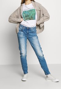 Vaqueros de denim azul claro con un ajuste entallado, textura desteñida y un ligero efecto de arrugado, combinados con una camiseta gráfica blanca y una camisa beige con estampado.