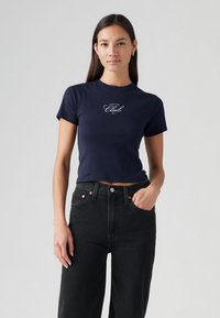 T-shirt azul-marinho curta em algodão com mangas curtas, apresentando a frase "Join the Club" impressa em letra branca cursiva. Combinada com jeans pretos de cintura alta.