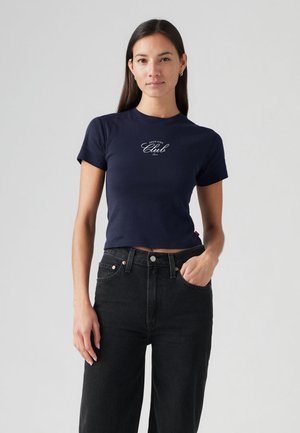 Īssis tumši zils T-krekls no kokvilnas ar īsām piedurknēm, uz kura baltā skripta veidā ir uzraksts "Nāc klāt kluba dalībniekam". Kombinēts ar melnām augsta jostasvietas džinsām.