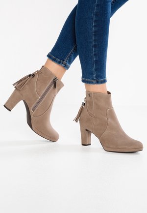Ben i blå jeans iført taupe ruskind ankellange støvler med sidelås og kvast, mellemhøj hæl, på et hvidt gulv.
