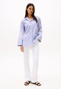 Camicia a righe blu e bianche con maniche lunghe, caratterizzata da ricami in pizzo sui polsini, abbinata a pantaloni bianchi e sandali neri.