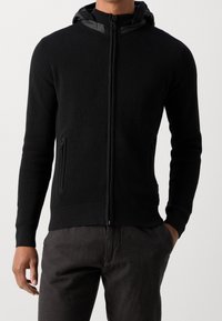 Black zip-up hoodie with ribbed texture, fitted design, and a contrasting smooth hood. Features side pockets and a snug waistband.