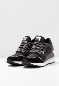 Para czarno-szarych butów sportowych z białymi podeszwami i wzorzystymi sznurówkami, umieszczonych pod lekkim kątem na białej powierzchni.