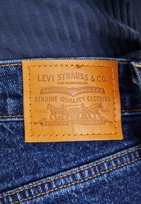 Nahka-Levi Strauss & Co. -tunnus ommeltuna sinisiin farkkuihin, jossa näkyvät brändin tiedot ja ommeltu oranssi lanka tumman kankaan päällä.