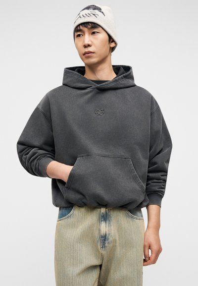 Γκρι φούτερ με κουκούλα και μπροστινή τσέπη, με λογότυπο στο στήθος, φορεμένο με ανοιχτόχρωμο, ξεβαμμένο φαρδύ τζιν. Το μοντέλο κοιτάζει ελαφρώς προς τα πάνω.