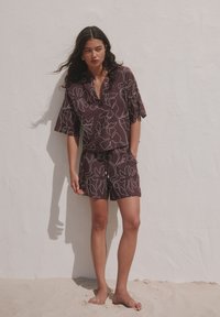 Jeune femme aux longs cheveux foncés, pieds nus sur le sable, portant une chemise à manches courtes et un short marron à motif floral assortis, devant un mur blanc.