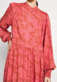 Abito lungo a maniche lunghe con motivo floreale rosa, collo alto, apertura frontale con bottoni, vita arricciata e polsini delle maniche elasticizzati, realizzato in un tessuto testurizzato.