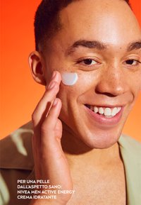 Una crema idratante viene applicata su una guancia. La persona ha la pelle chiara con lentiggini, capelli corti, denti bianchi e indossa una maglietta verde chiaro. Sfondo arancione.