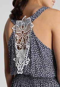 Námořnické květinové šaty s bílými krajkovými detaily na zádech. Mají bezramenný design, složitý krajkový vzor a lehký materiál.