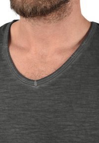 Tricou din bumbac gri cu decolteu în V, având o textură subtilă și o croială lejeră, prezentat până la linia gâtului.