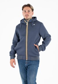Sweatshirt zippé gris avec capuche, featuring une bande jaune et blanche sur le devant, des poignets côtelés et un petit logo sur la poitrine.
