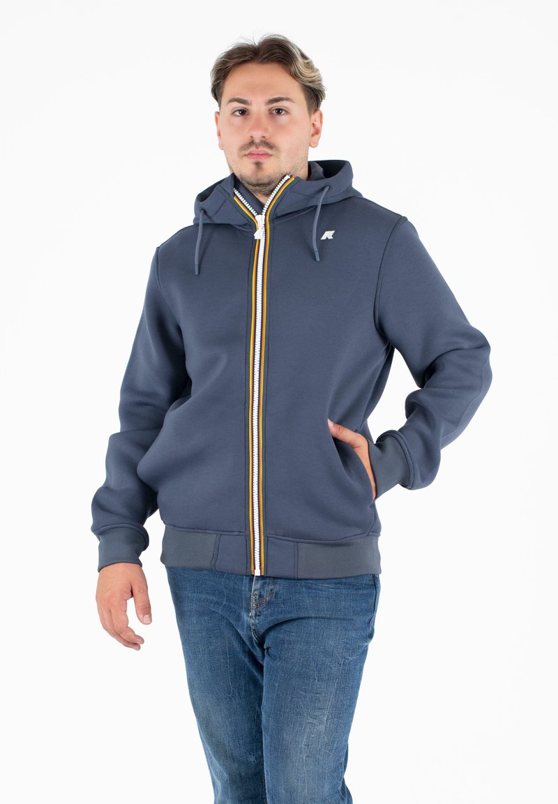 Sweatshirt zippé gris avec capuche, featuring une bande jaune et blanche sur le devant, des poignets côtelés et un petit logo sur la poitrine.