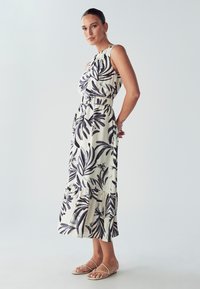Maksi obleka z belo tkanino in velikimi črnimi tropskimi listnimi vzorci, elastičnim pasom in volanastim robom. v kombinaciji s sandalami brez pet.