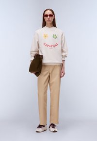 Φούτερ σε κρεμ χρώμα με γραφικά λουλουδιών, συνδυασμένο με μπεζ παντελόνια και λευκά αθλητικά παπούτσια με μαρόν λεπτομέρειες. Φοράει καφέ τσάντα.