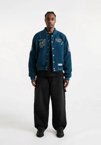 Man in een blauwe bomberjack met patches, zwarte wijde broek en zwarte schoenen, staande tegen een effen witte achtergrond.