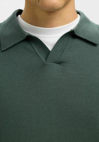 Zeleni polo majica s teksturom, s ovratnikom i V-izrezom, prebacena preko bijele majice s okruglim izrezom.
