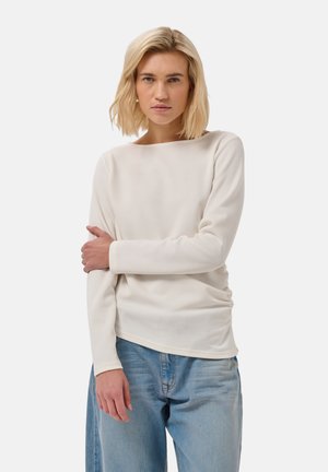 Femme aux cheveux blonds portant un haut blanc à manches longues et un jean bleu clair, debout devant un fond blanc uni, regardant devant elle.