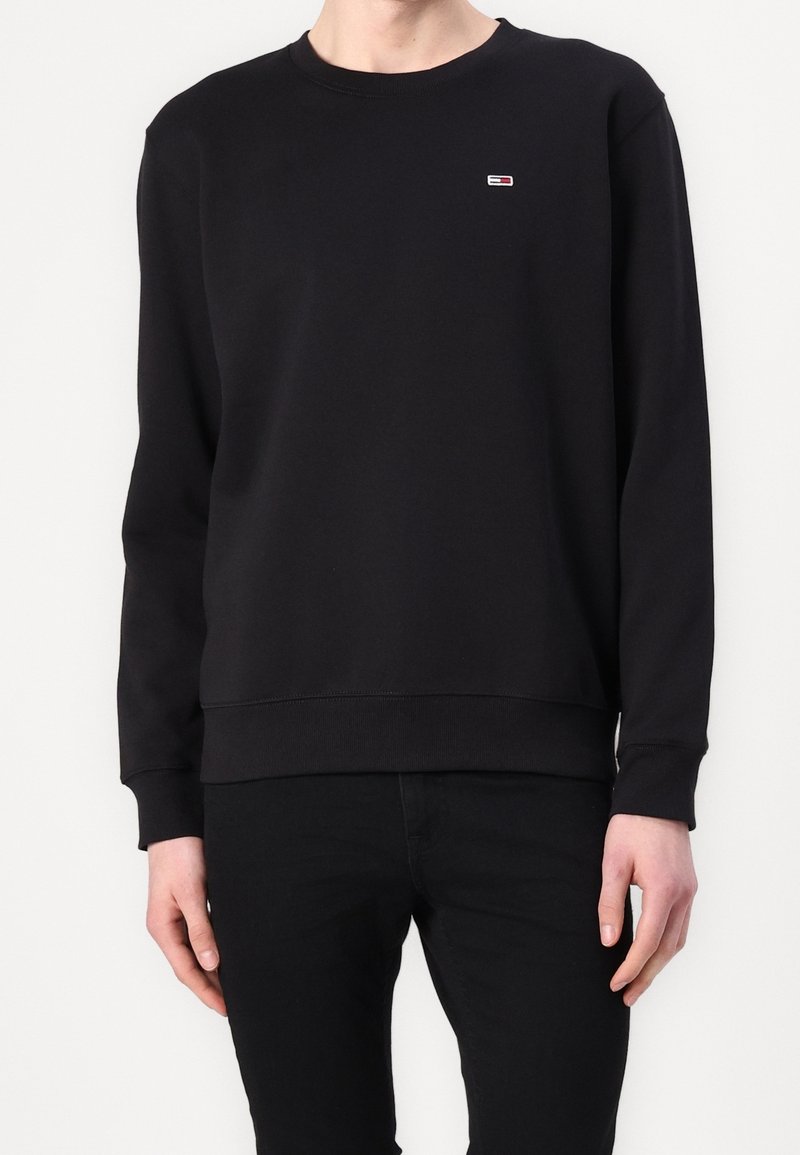 Personne portant un sweat-shirt noir uni à manches longues avec un petit logo rectangulaire sur la poitrine gauche, associé à un pantalon noir.