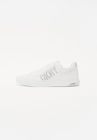 White sneakers featuring a smooth leather upper, flat sole, lace-up front, and a sparkling "DKNY" logo accent on the side.