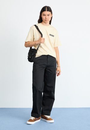 Beige short-sleeve t-shirt with "DICKIES" logo, black wide-leg trousers, and brown sneakers with white soles. Small black crossbody bag.