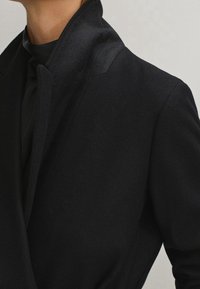Veste noire au design épuré. Dotée d'un col satin et d'un tissu doux et texturé. Détail de couture latérale visible.